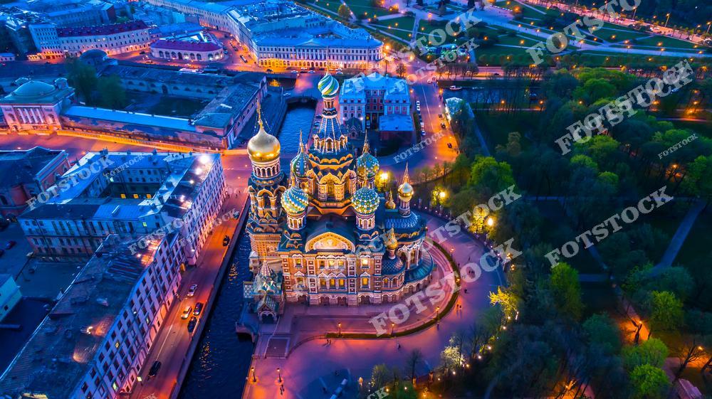 На фотографии изображён храм Спас-на-Крови, расположенный на набережной канала Грибоедова, одна из самых знаковых достопримечательностей Санкт-Петербурга. Его яркие купола и детализированные фасады подчёркнуты вечерней подсветкой, создающей сказочную атмосферу. Вокруг видны аллеи и зелёные зоны Летнего сада, а свет уличных фонарей усиливает ощущение уюта и величия архитектуры. Вид с высоты даёт возможность оценить красоту этого уголка Петербурга в ночное время.